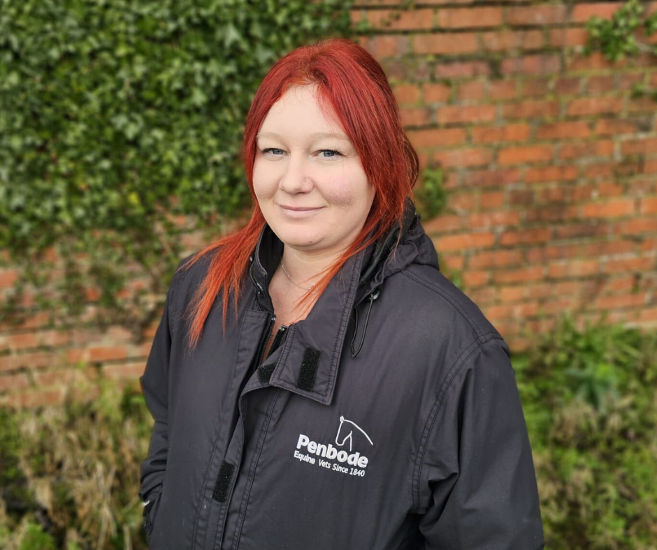 A woman with long red hair wearing a black Penbode Equine Vets jacket stands outdoors in front of a brick wall and green foliage, smiling gently at the camera.