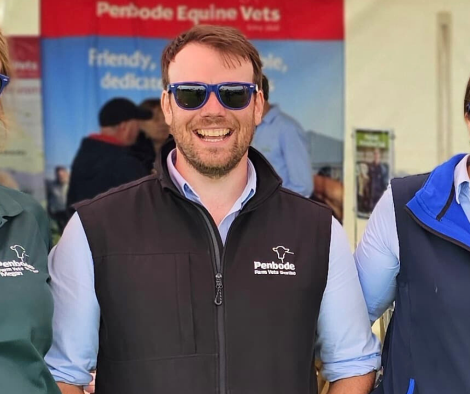 A smiling man wearing sunglasses and a black Penbode Farm Vets vest stands with two colleagues, with a Penbode Equine Vets display banner in the blurred background.