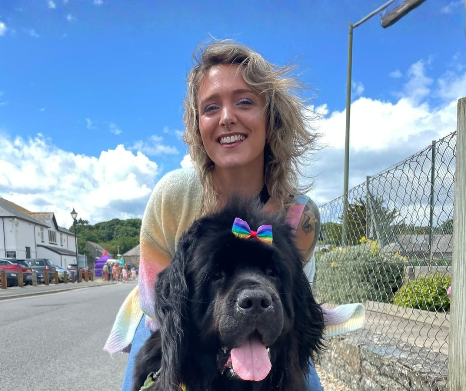 A smiling woman in a pastel sweater poses outdoors with a large black dog wearing a rainbow bow on its head. The sky is blue with scattered clouds and there are houses and cars along the street.