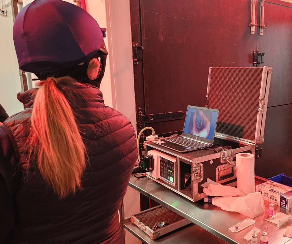 A person wearing a helmet and jacket stands near a metal table, watching a laptop displaying a medical image. Medical equipment, gloves, and paper towels are arranged nearby. The scene is lit by red and white lights.